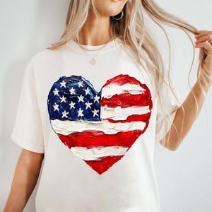 Pu&ograve; includere: Maglietta bianca con una bandiera americana dipinta a forma di cuore.