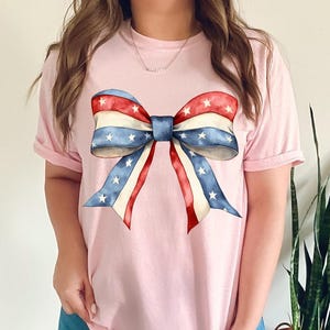 K&ouml;nnte beinhalten: Ein pinkfarbenes T-Shirt mit einer gro&szlig;en rot-wei&szlig;-blauen Schleife mit Sternen darauf.