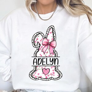 Pu&ograve; includere: Felpa bianca con un design di coniglio floreale rosa e il nome "Adelyn" stampato sotto. Il coniglio ha un fiocco rosa e un cuore rosa al centro.