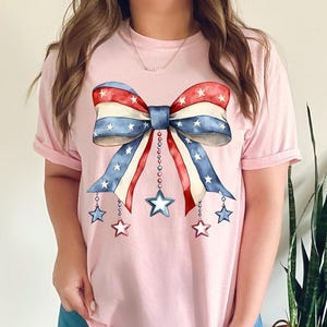 May include: A pink t-shirt with a red, white, and blue bow with stars and chains hanging from it. The bow is in the style of the American flag.