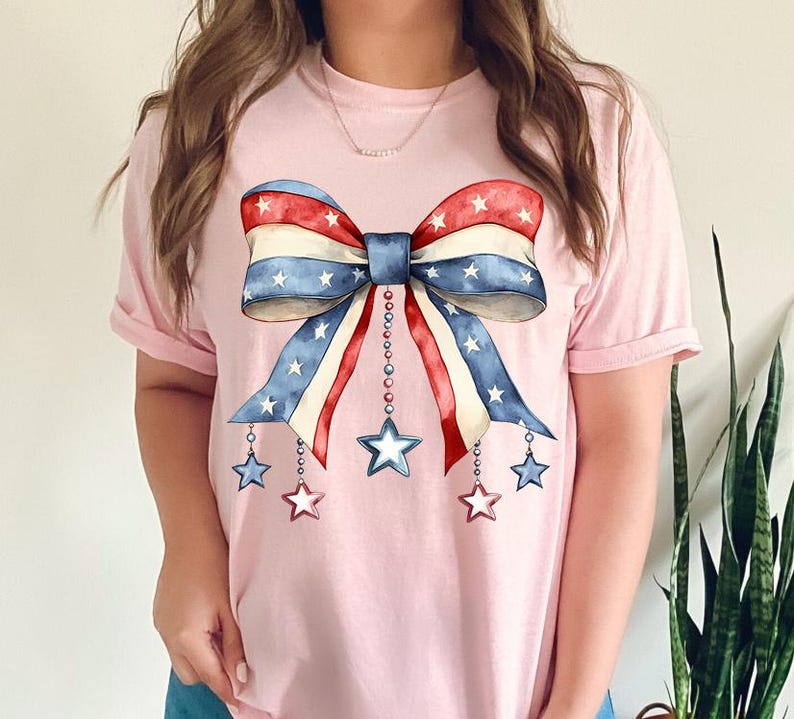 May include: A pink t-shirt with a red, white, and blue bow with stars and chains hanging from it. The bow is in the style of the American flag.