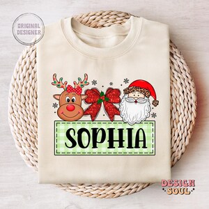 May include: A beige sweatshirt with a green and white checkered banner that says "Sophia". The banner is flanked by a cartoon reindeer with a red nose and a cartoon Santa Claus with a white beard. The reindeer and Santa Claus are surrounded by red and white Christmas decorations.