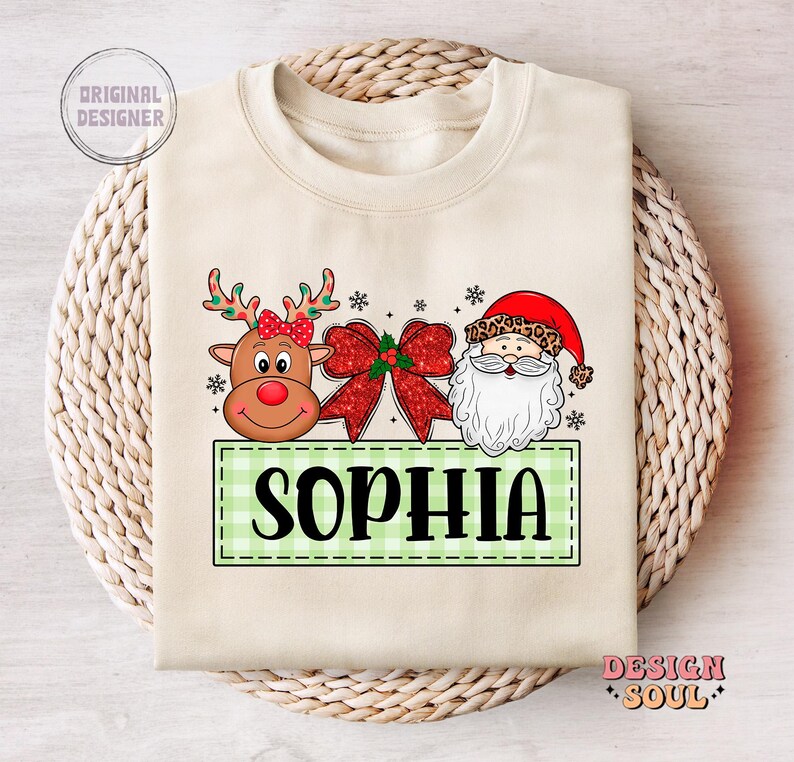 May include: A beige sweatshirt with a green and white checkered banner that says "Sophia". The banner is flanked by a cartoon reindeer with a red nose and a cartoon Santa Claus with a white beard. The reindeer and Santa Claus are surrounded by red and white Christmas decorations.