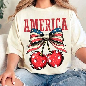 May include: A white t-shirt with the word "AMERICA" printed in red, with a red, white, and blue bow and two cherries with white stars printed below the word.