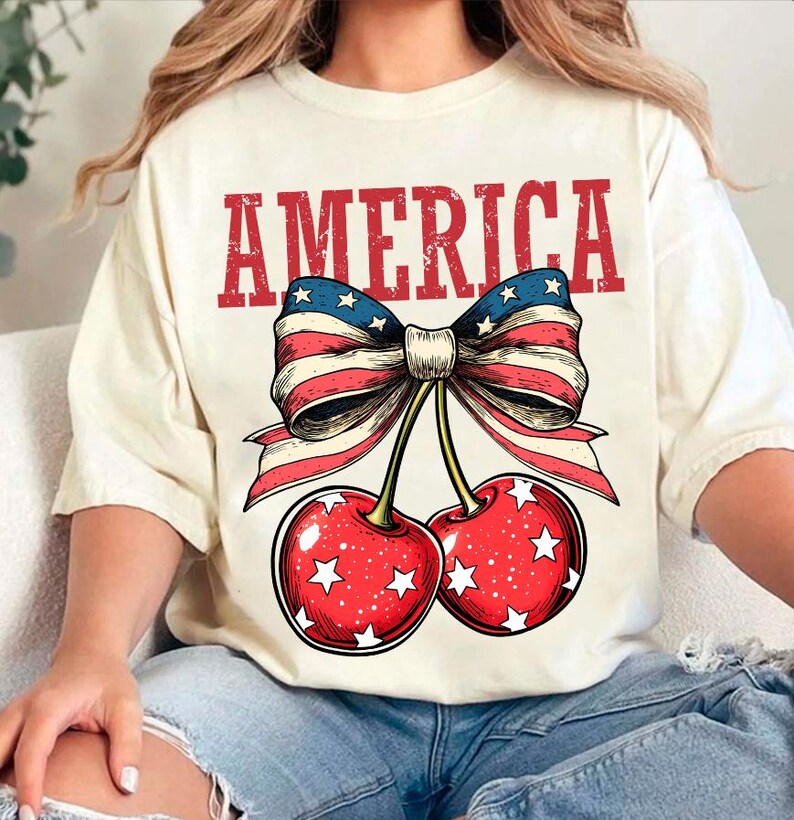 May include: A white t-shirt with the word "AMERICA" printed in red, with a red, white, and blue bow and two cherries with white stars printed below the word.
