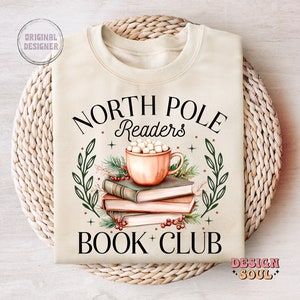 May include: A beige sweatshirt with the text "North Pole Readers Book Club" in black. The design features a mug of hot chocolate with marshmallows, a stack of books, and green leaves.