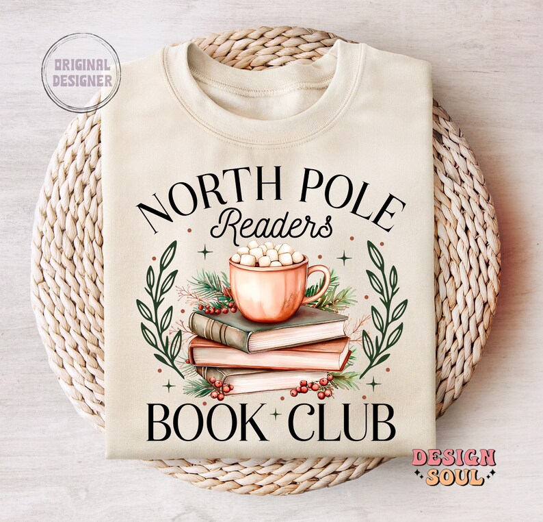 May include: A beige sweatshirt with the text "North Pole Readers Book Club" in black. The design features a mug of hot chocolate with marshmallows, a stack of books, and green leaves.
