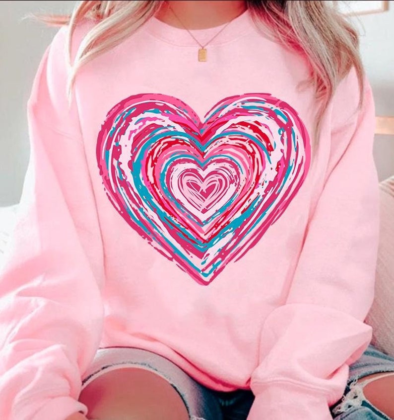 Pu&ograve; includere: Felpa rosa con un grande disegno a forma di cuore colorato. Il cuore &egrave; composto da cerchi concentrici di vernice rosa, blu e bianca.
