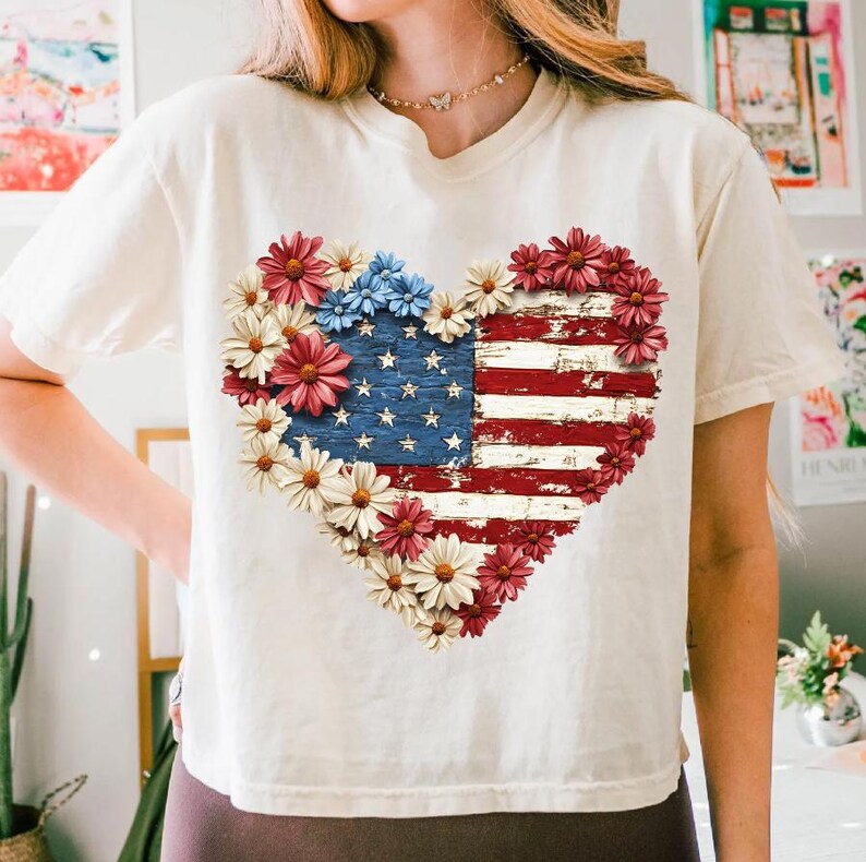 May include: Cream-colored t-shirt featuring a heart-shaped design of the American flag. The flag is made of distressed wood and surrounded by red, white, and blue flowers. The shirt has short sleeves and a crew neck.