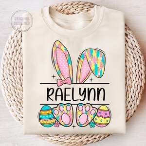 May include: A light beige sweatshirt with a personalized Easter bunny design. The bunny ears are pink and blue with white polka dots and gold glitter. The name "Raelynn" is printed in black below the bunny ears. The design also includes two pink paw prints and three colorful Easter eggs.