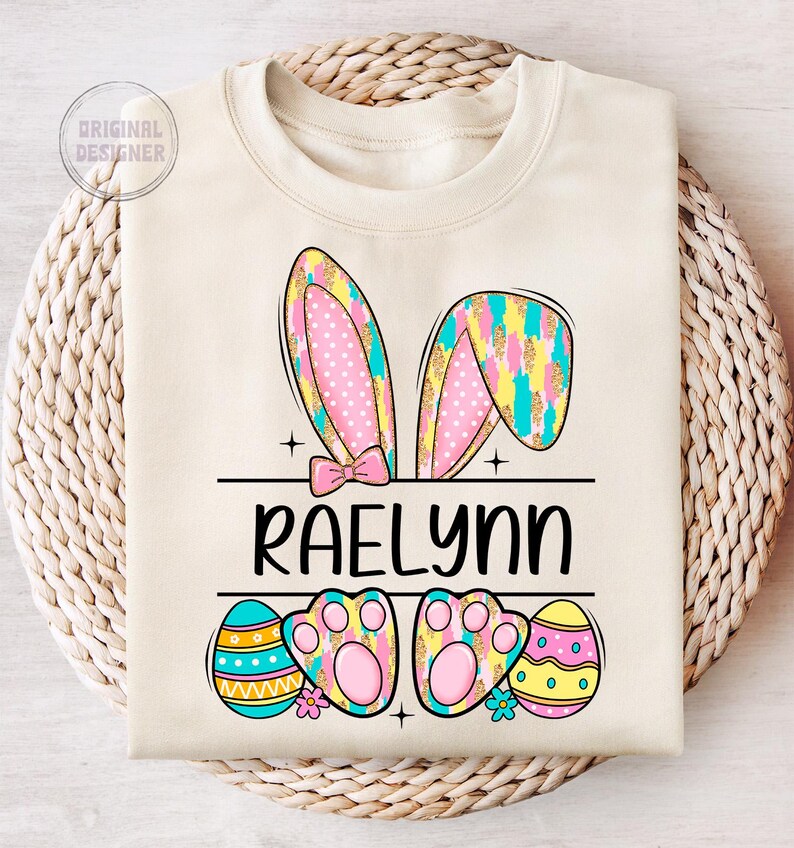 May include: A light beige sweatshirt with a personalized Easter bunny design. The bunny ears are pink and blue with white polka dots and gold glitter. The name "Raelynn" is printed in black below the bunny ears. The design also includes two pink paw prints and three colorful Easter eggs.