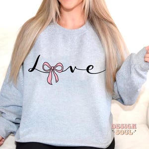 May include: A light gray sweatshirt with the word "love" written in black cursive with a pink bow in the middle of the word.