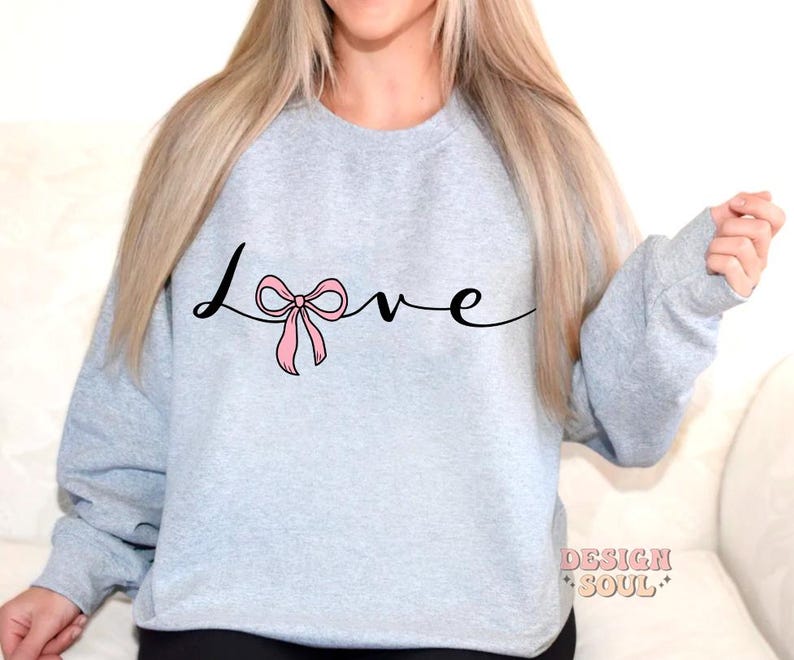 May include: A light gray sweatshirt with the word "love" written in black cursive with a pink bow in the middle of the word.