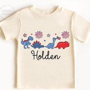 May include: Cream-colored t-shirt featuring a row of cartoon dinosaurs in blue and red, with fireworks and stars. The name "Holden" is written in black script below the dinosaurs. The shirt has short sleeves.