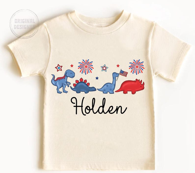 May include: Cream-colored t-shirt featuring a row of cartoon dinosaurs in blue and red, with fireworks and stars. The name "Holden" is written in black script below the dinosaurs. The shirt has short sleeves.
