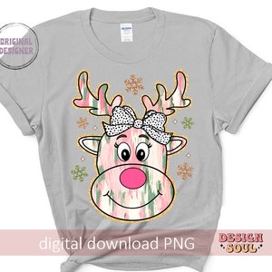May include: A gray t-shirt with a pink and green reindeer with a pink bow and black polka dots. The reindeer has a pink nose and a white smile. The background is white with gold snowflakes.