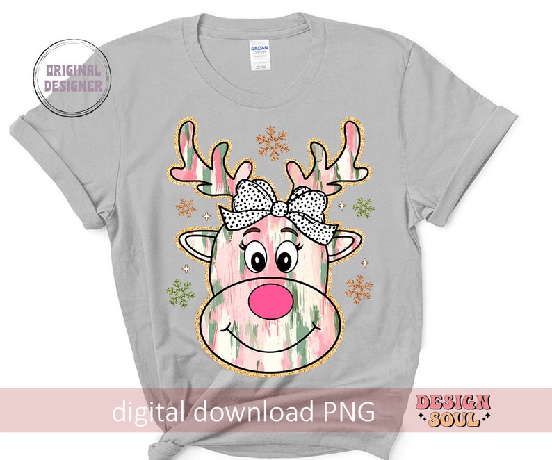 May include: A gray t-shirt with a pink and green reindeer with a pink bow and black polka dots. The reindeer has a pink nose and a white smile. The background is white with gold snowflakes.