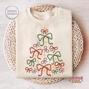 May include: A cream-coloured sweatshirt featuring a Christmas tree design made from green, red and pink bows. The bows are arranged in a tree shape with yellow stars and red dots.