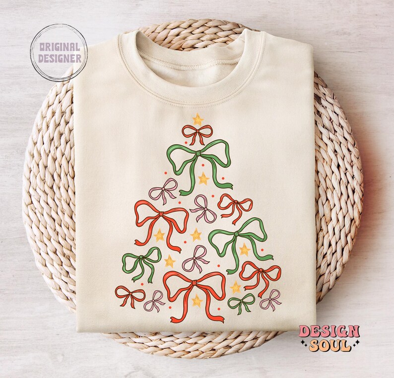 May include: A cream-coloured sweatshirt featuring a Christmas tree design made from green, red and pink bows. The bows are arranged in a tree shape with yellow stars and red dots.