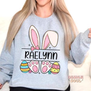 May include: A light gray sweatshirt with a bunny ear design, the name "Raelynn" printed in black, and colorful Easter eggs.