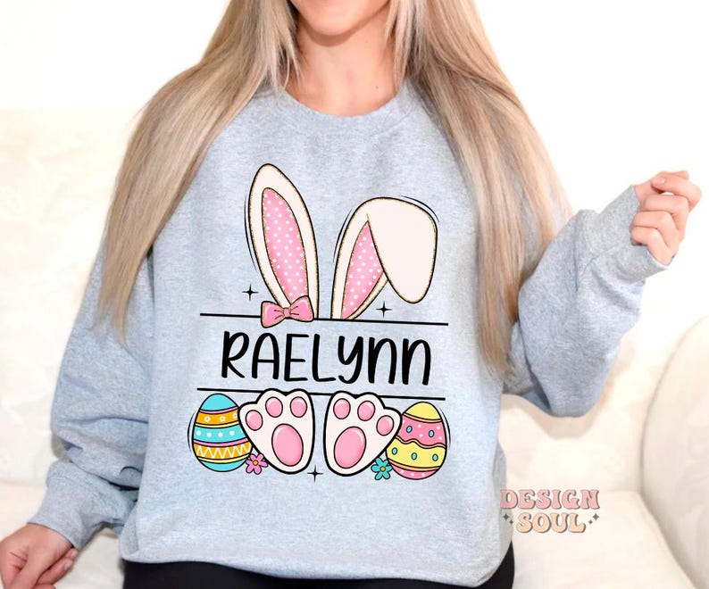 May include: A light gray sweatshirt with a bunny ear design, the name "Raelynn" printed in black, and colorful Easter eggs.