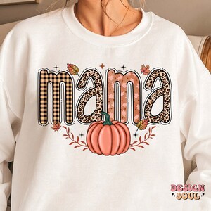 May include: White sweatshirt with a graphic design of the word "mama" in different patterned letters, including plaid, leopard print, and floral. A small pumpkin is below the word.