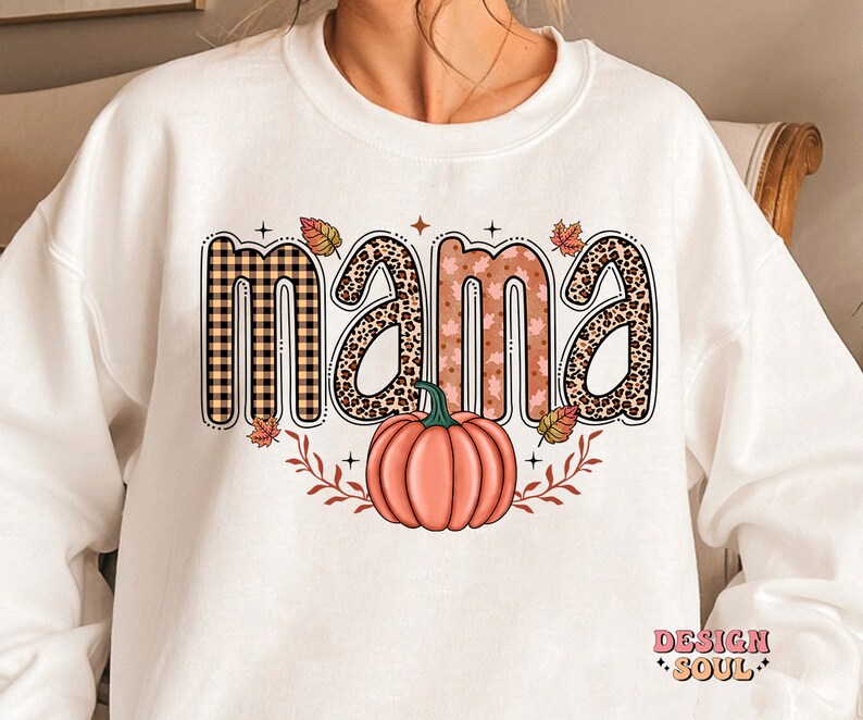 May include: White sweatshirt with a graphic design of the word "mama" in different patterned letters, including plaid, leopard print, and floral. A small pumpkin is below the word.