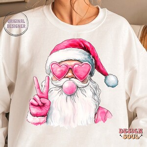 May include: White sweatshirt featuring a pink and white illustration of Santa Claus wearing pink heart-shaped sunglasses and blowing a pink bubble. Santa is giving the peace sign with his right hand. The text "Design Soul" is printed in black at the bottom right corner of the sweatshirt.
