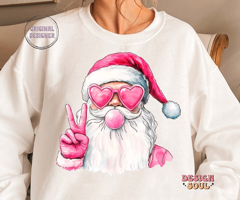 May include: White sweatshirt featuring a pink and white illustration of Santa Claus wearing pink heart-shaped sunglasses and blowing a pink bubble. Santa is giving the peace sign with his right hand. The text "Design Soul" is printed in black at the bottom right corner of the sweatshirt.