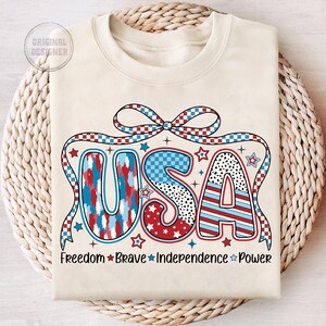 Peut inclure: Un sweat-shirt beige avec un graphique USA rouge, blanc et bleu. Le graphique est compos&eacute; d'&eacute;toiles, de rayures et de motifs &agrave; carreaux. Les mots "Freedom Brave Independence Power" sont imprim&eacute;s sous le graphique.