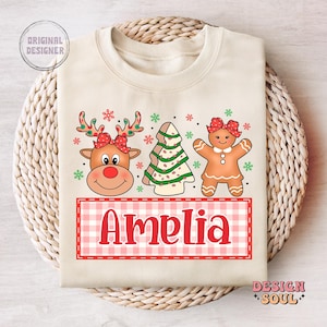 May include: A cream-colored sweatshirt with a Christmas-themed design featuring a gingerbread person, a reindeer, and a Christmas tree. The text "Amelia" is printed in red on a pink and white checkered background.
