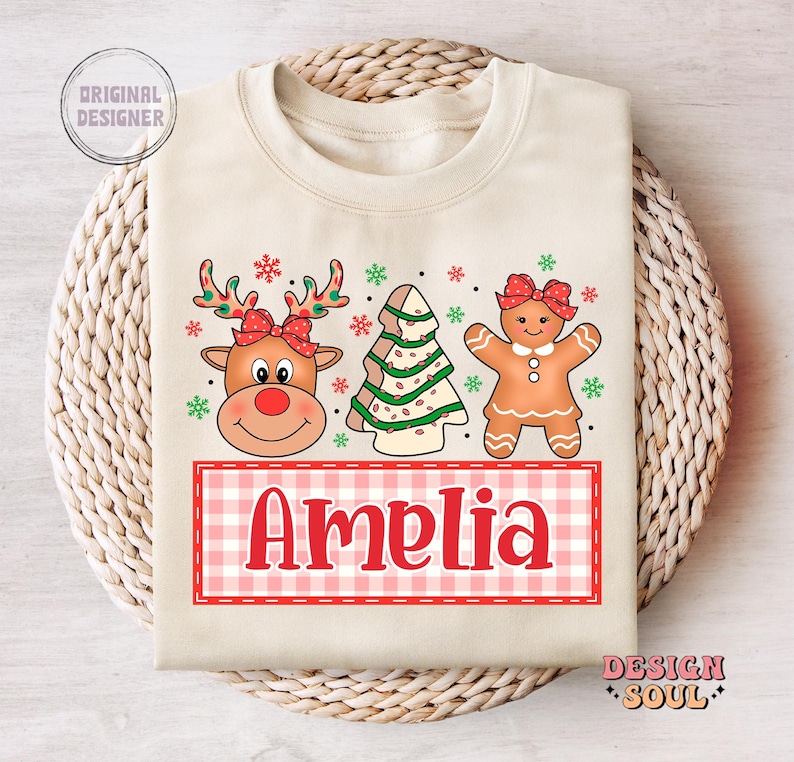May include: A cream-colored sweatshirt with a Christmas-themed design featuring a gingerbread person, a reindeer, and a Christmas tree. The text "Amelia" is printed in red on a pink and white checkered background.