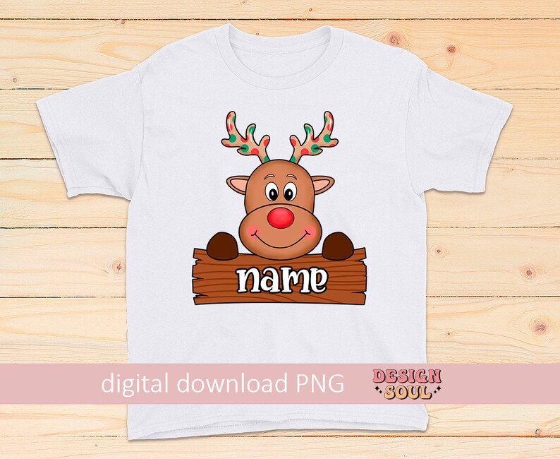 Peut inclure: T-shirt blanc avec un renne de dessin anim&eacute; tenant une pancarte qui dit "name". Le renne a des bois rouges avec des pois verts et blancs.