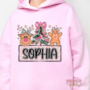 Op de afbeelding: Roze hoodie met een kerstthema met een gingerbread man, een rendier en een kerstboom. Het ontwerp bevat de naam "Sophia".