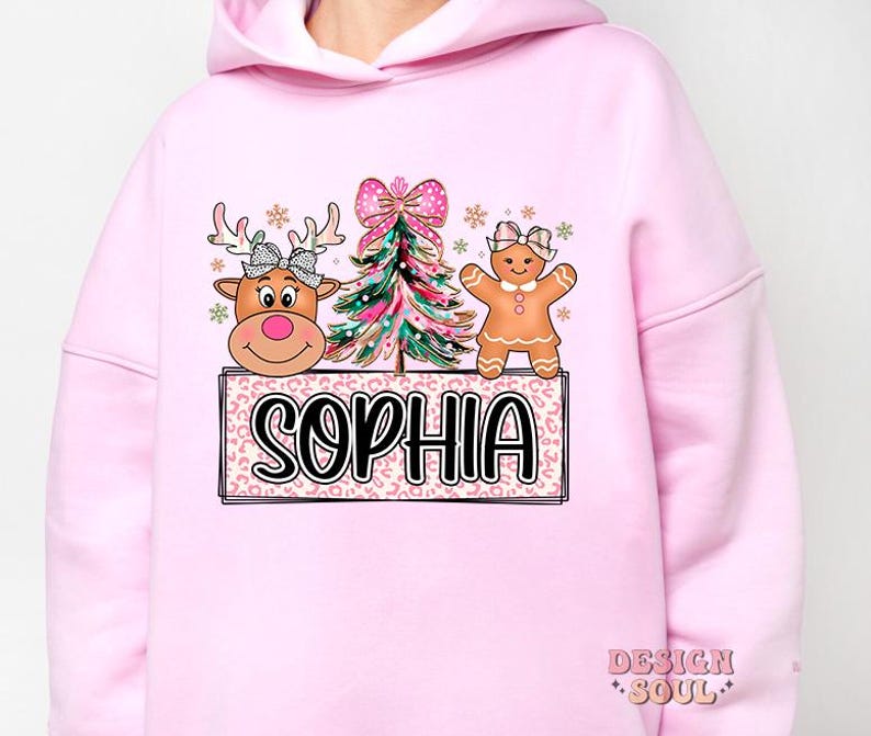 Op de afbeelding: Roze hoodie met een kerstthema met een gingerbread man, een rendier en een kerstboom. Het ontwerp bevat de naam "Sophia".