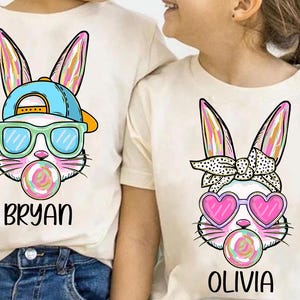 May include: Two white t-shirts with a cartoon bunny design. One bunny is wearing sunglasses and a blue baseball cap and has the name "Bryan" printed below. The other bunny is wearing pink heart-shaped sunglasses and a polka dot headband and has the name "Olivia" printed below.