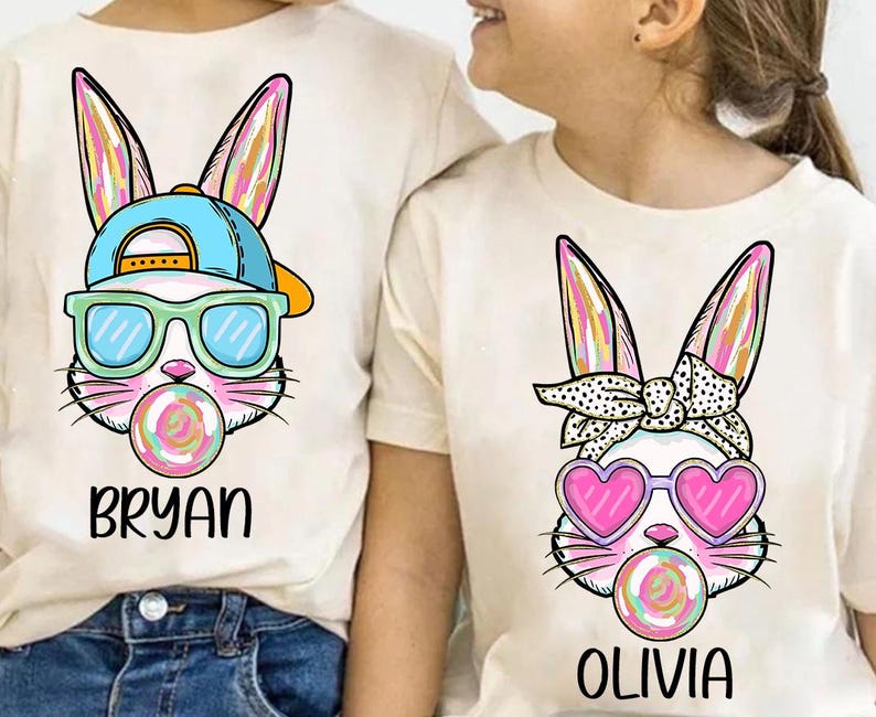 May include: Two white t-shirts with a cartoon bunny design. One bunny is wearing sunglasses and a blue baseball cap and has the name "Bryan" printed below. The other bunny is wearing pink heart-shaped sunglasses and a polka dot headband and has the name "Olivia" printed below.