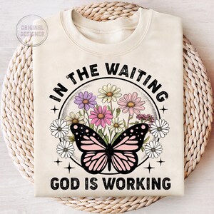 May include: A beige t-shirt with a black butterfly and floral design. The text "In the waiting God is working" is printed on the shirt.