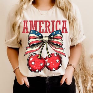 May include: A white t-shirt with a red, white, and blue bow with cherries and the word "AMERICA" printed on it.