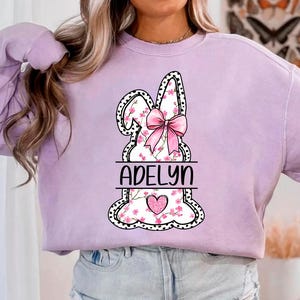 Pu&ograve; includere: Una felpa color lavanda con una grafica di un coniglio rosa. Il coniglio ha un fiocco rosa e dettagli floreali. Il nome "Adelyn" &egrave; stampato sotto il coniglio.