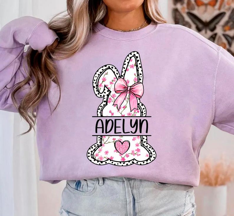 Pu&ograve; includere: Una felpa color lavanda con una grafica di un coniglio rosa. Il coniglio ha un fiocco rosa e dettagli floreali. Il nome "Adelyn" &egrave; stampato sotto il coniglio.