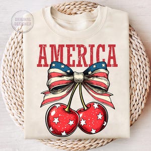 May include: A white t-shirt with the word "AMERICA" printed in red letters. A red, white, and blue bow with stars is tied around two red cherries with white stars.