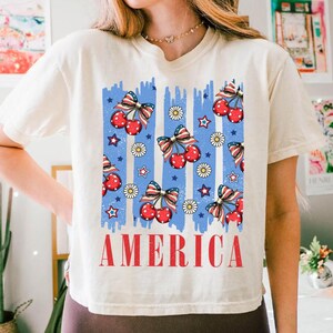 May include: A white t-shirt with a blue and red graphic print of cherries, stars, and daisies. The graphic is reminiscent of an American flag. The text "AMERICA" is printed in red below the graphic.