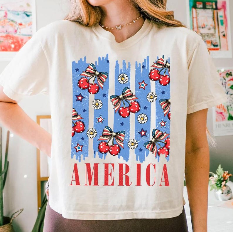 May include: A white t-shirt with a blue and red graphic print of cherries, stars, and daisies. The graphic is reminiscent of an American flag. The text "AMERICA" is printed in red below the graphic.