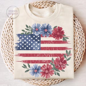 Puede incluir: Sudadera color crema con un dise&ntilde;o de bandera estadounidense desgastada con detalles florales rojos y azules. El dise&ntilde;o de la bandera incluye estrellas y rayas, con una corona floral alrededor de la bandera. El texto "ORIGINAL DESIGNER" es visible.