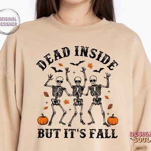May include: Beige sweatshirt with the text "DEAD INSIDE BUT IT'S FALL" and a graphic of three dancing skeletons wearing sunglasses. The design includes bats, leaves, and pumpkins, perfect for Halloween or autumn.
