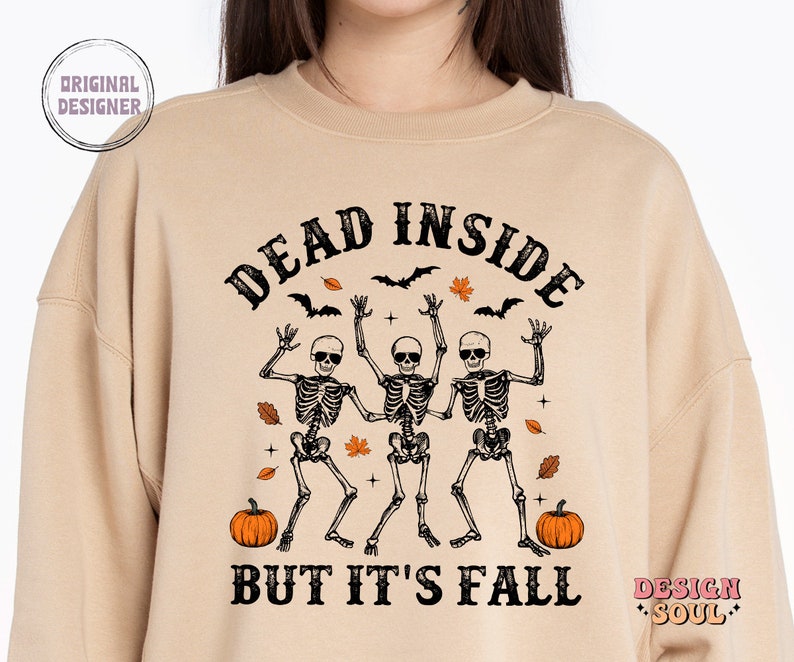 May include: Beige sweatshirt with the text "DEAD INSIDE BUT IT'S FALL" and a graphic of three dancing skeletons wearing sunglasses. The design includes bats, leaves, and pumpkins, perfect for Halloween or autumn.