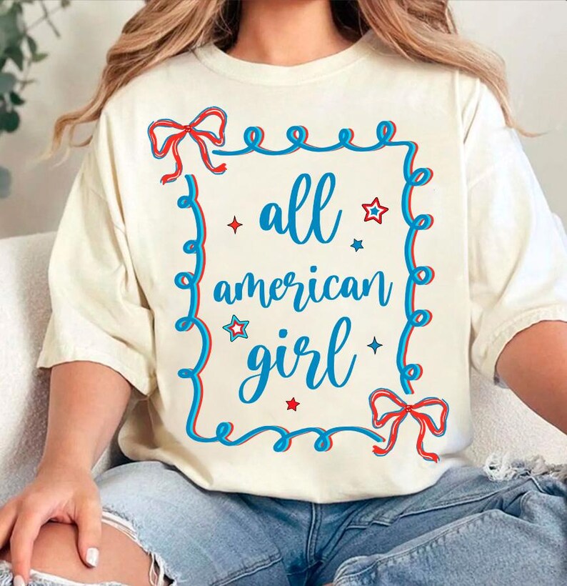 Peut inclure: Un t-shirt blanc avec un motif bleu et rouge qui dit "all american girl" &agrave; l'int&eacute;rieur d'un cadre bleu avec des &eacute;toiles rouges et des n&oelig;uds.