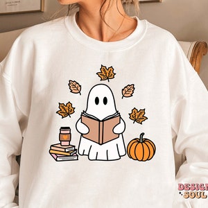 May include: White sweatshirt with a cute ghost reading a book surrounded by autumn leaves. The ghost is wearing a pink heart. The sweatshirt also has a cup of coffee and a stack of books. The text "DESIGN SOUL" is in the bottom right corner.