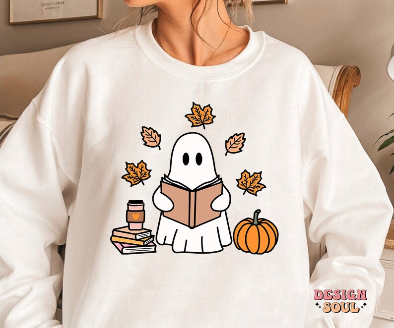 May include: White sweatshirt with a cute ghost reading a book surrounded by autumn leaves. The ghost is wearing a pink heart. The sweatshirt also has a cup of coffee and a stack of books. The text "DESIGN SOUL" is in the bottom right corner.